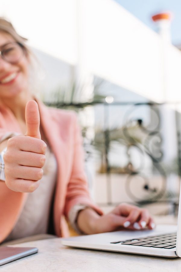 Cute young woman, student, business lady showing thumbs up, well done, sitting in outdoor cafe on terrace with laptop. Wearing pink smart clothes.